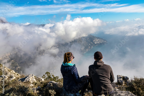 Happy couple with panoramic view from Monte Comune on the coastal town Positano appearing from clouds. Magical hiking above fog in Lattari Mountains, Apennines, Amalfi Coast, Campania, Italy, Europe