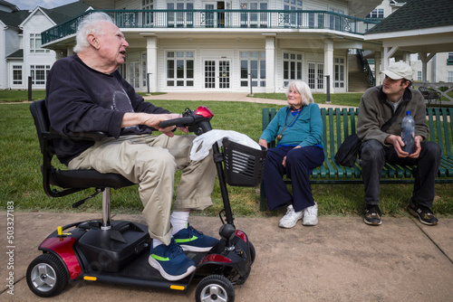 Solomons, Maryland A senior man sits outdoors on an electric wheelchair and chats with his wife and grandson.