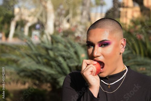Portrait of non-binary person, young and South American, heavily make up, biting a red apple that she holds in her hand and surrounded by vegetation. Concept queen, lgbtq+, pride, queer, sin.