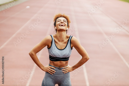A happy runner stands in the stadium with her hands on hips and laughing. She is so happy she accomplished her goal and moved boundaries. A runner resting and laughing.