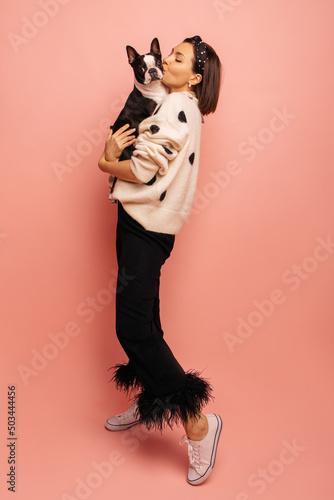 Full length young caucasian woman kissing her black and white dog while standing on pink background. Brunette with bob haircut wears sweater and pants. True friendship, wonderful happy moments