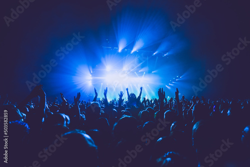 A crowded concert hall with scene stage lights in blue tones, rock show performance, with people silhouette, on dance floor air during a concert festival