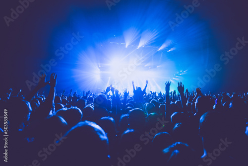 A crowded concert hall with scene stage lights in blue tones, rock show performance, with people silhouette, on dance floor air during a concert festival