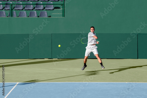 Professional tennis player performs forehand hit on hard tennis court. Young male athlete with tennis racket in action. Junior tennis sport. Green background, banner, copy space