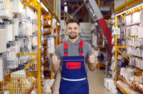 Happy salesman at modern DIY store guarantees best quality of all the goods. Portrait of handsome young man in uniform standing between shelves with ventilation grids, smiling and showing thumbs up