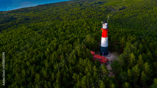 Bird's eye view of the lighthouse surrounded by the green forest in a daylight. Sea in the distance.