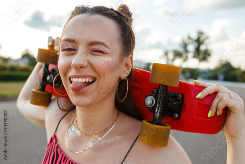 Positive young teenage female skater with skateboard showing tongue and making funny face while looking at camera in city street against sunset sky in summer 