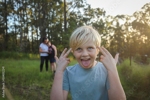Blonde caucasian boy making silly face expression in the bush with mother and brother hugging out of focus in background