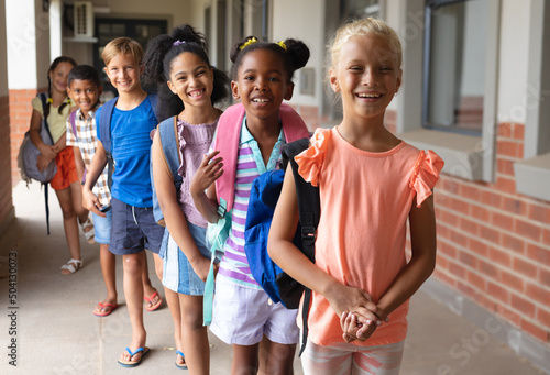 Portrait of smiling multiracial elementary school students with backpack standing in row at school