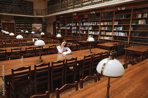 Female student sitting alone in a public old library and reading a book with a serious face.