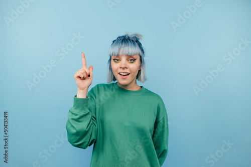 Positive lady with blue hair stands on a blue background with a happy face looking at the camera and showing finger up