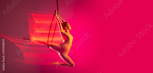 Dynamic portrait of young artistic girl, professional air gymnast training, performing isolated over pink background with mixed lights effect. Flyer