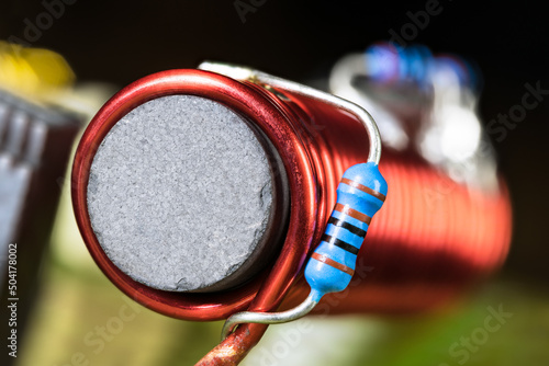 Cylindrical inductor with black ferromagnetic core wrapped by red wire on PCB detail. Closeup of small blue carbon resistors with electronic color code soldered on coil winding with blurry background.