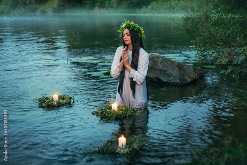 Slavic woman holds herbal wreath in hands puts on water, candles float. Fantasy girl nymph White long dress. Summer green grass tree river bank. Concept pagan holiday of Ivan Kupala divination maidens