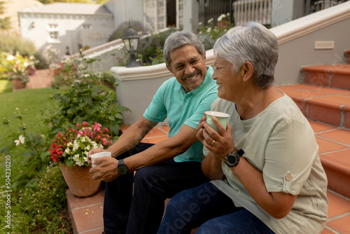 Smiling biracial senior man talking with senior wife holding coffee cup while sitting on steps