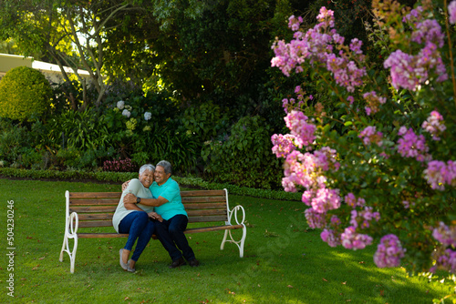 Romantic biracial senior man embracing wife while sitting on bench against trees and plants in park