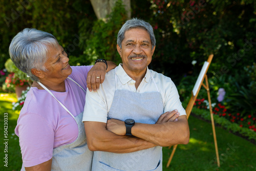 Smiling biracial senior woman with hand on husband's shoulder standing against plants in yard
