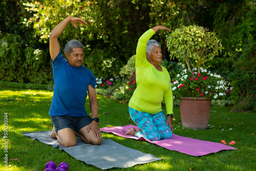Biracial senior couple with arm raised kneeling on yoga mats while exercising against plants in yard