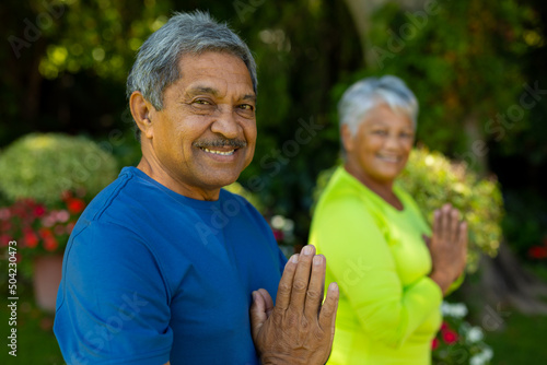 Portrait of smiling biracial senior couple standing in prayer position against plants in yard