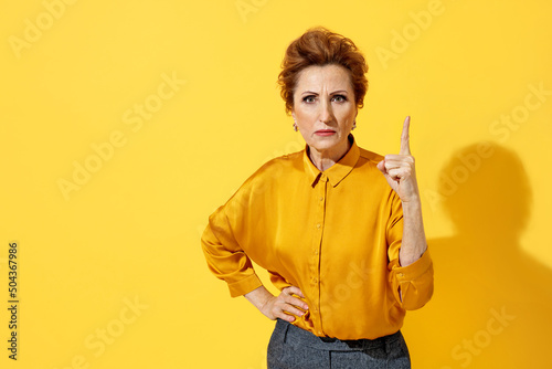 Elderly strict woman point index finger up, warns, notifies, recommends something. Photo of attractive woman in yellow shirt on yellow background