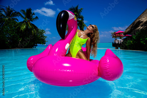 Beautiful sexy tanned woman on pink flamingo in pool. Young glamour girl in swimsuit on Maldives island. Perfect body bikini model in luxury resort on Maldives. Luxury travel. Summertime.