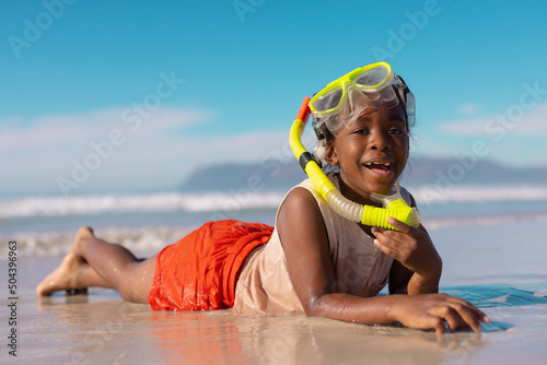 Portrait of happy african american wet girl wearing mask and snorkel lying at shore against blue sky