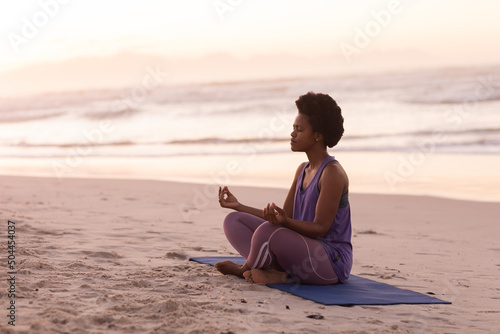 African american mature woman meditating while sitting on mat against sea and clear sky at sunset