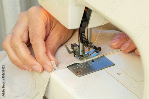 A woman sews on a sewing machine. Women's hands substitute the fabric for sewing under the foot of the sewing machine.