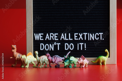 Toy dinosaurs next to a felt board that says We are all extinct. Don't do it! Red background. Concept agitating against self-destruction and extinction of mankind.