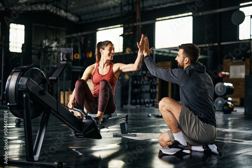 Happy athlete giving high-five to her personal trainer after exercising on rowing machine at gym.