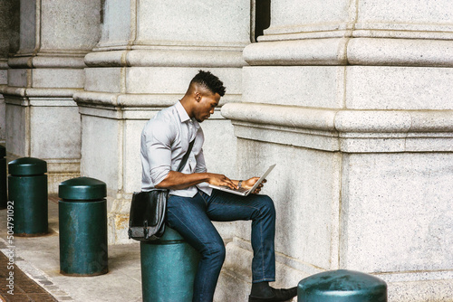 African American College Student studying in New York, wearing shirt, jeans, carrying shoulder leather bag, sitting on pillar on street, reading, working on laptop computer. filtered effect..