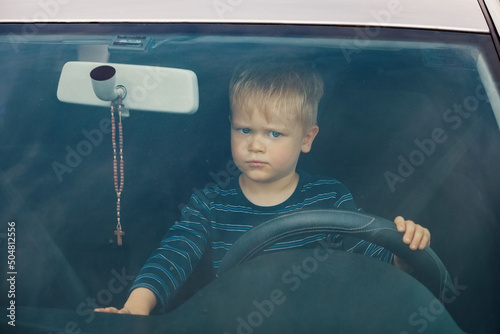 Portrait of a child sitting in a car behind the wheel from the front. The boy plays and imagines he is driving, he controls the radio with the other hand.