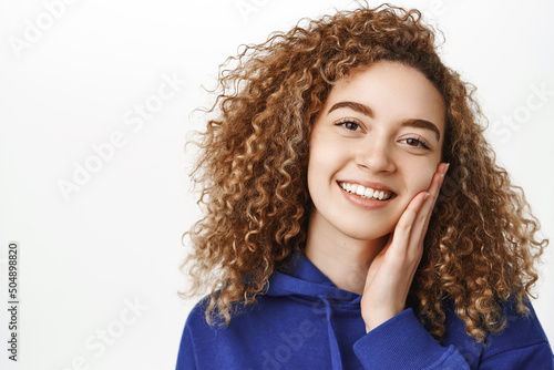 Beauty and wellbeing. Close up portrait of beautiful curly girl with perfect, healthy face, touching her skin and smiling pleased, standing over white background