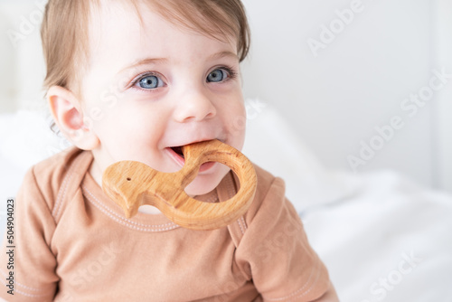 portrait of baby girl with blue eyes plays with wooden toy teether for teeth on white background