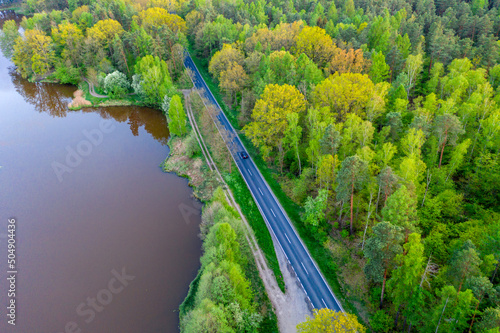 Zbiornik wodny, jezioro położone w lesie. Brzegi porośnięte drzewami. Obok przebiega asfaltowa droga. Zdjęcie z drona.