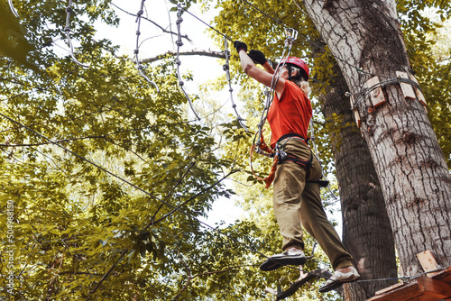 A woman climbs on a tourist rope town, which is located high among the trees, in a climbing harness, fastened with a carabiner belay, on the head of a helmet.