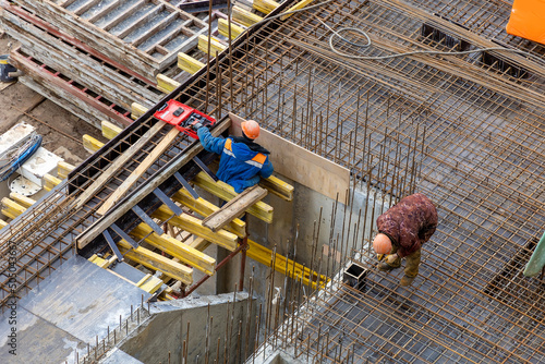 Builders in orange helmets make a steel reinforcement cage for pouring a concrete slab. Steel rods and metal formwork on a construction site. Monolithic construction. Cast-in-place concrete