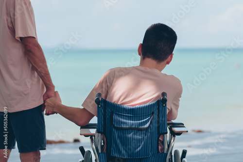 Wheelchair and behind of young man and father's, Dad hand holding the son's hand staring at the sea ahead,Family vacation activity,Encourage to strengthen,Mental health and travel or vacation concept.