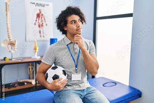 Hispanic man with curly hair working as football physiotherapist with hand on chin thinking about question, pensive expression. smiling with thoughtful face. doubt concept.