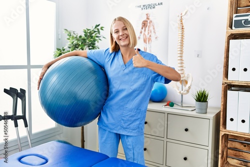 Young caucasian physiotherapist woman holding pilates ball smiling happy and positive, thumb up doing excellent and approval sign
