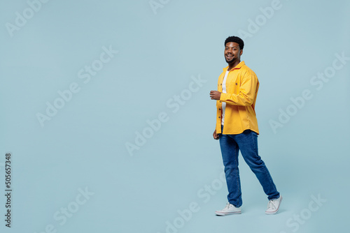 Full body young man of African American ethnicity 20s wear yellow shirt walking going strolling look camera isolated on plain pastel light blue background studio portrait. People lifestyle concept