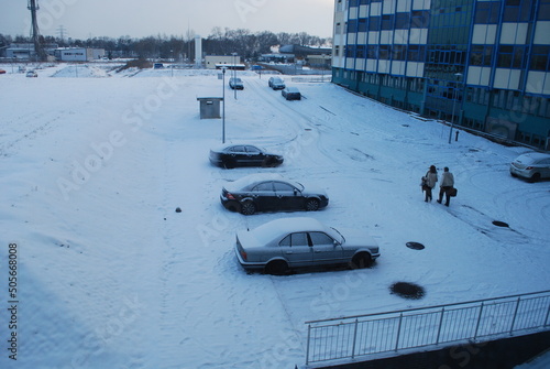 view of the building wall and parking and cars lot in winter, snow, sunlight breaking through the clouds