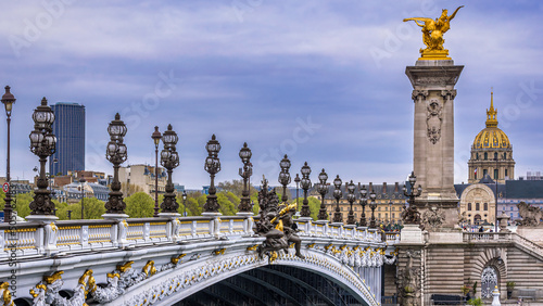 The ornate Pont Alexandre III in the downtown of Paris, France with the golden dome of the Invalides and the Tour Montparnasse in the background below an overcast sky