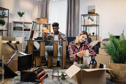 Positive woman unpacking boxes and talking on mobile phone while man assembling furniture behind. Young multiracial couple enjoying moving process to new house.