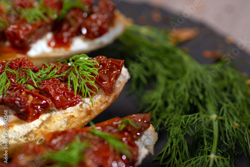 Antipasti - bruschetta with the creamy cheese, dried tomatoes and the dill on the black stone plate on a blurred background, closeup.