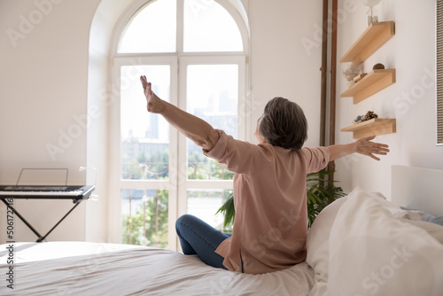 Carefree senior lady feeling full of energy after sleep enough, stretching body, opening hands, looking at window. Middle aged grey haired mature woman sitting in beddings, on comfortable mattress
