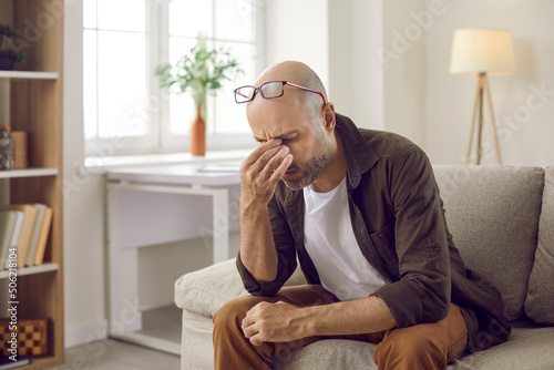 Unhealthy man feel stressed suffer from migraine or headache at home. Unwell tired middle-aged male sit on sofa struggle with blurry vision or dizziness, have high blood pressure. Health problem.