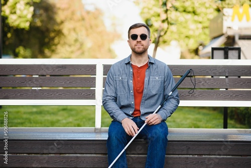 Visually impaired man with walking stick, sitting on bench in city park. Copy space