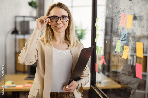 Smiling business lady with clipboard standing near office glass wall with sticky notes and looking at camera. Female entrepreneur wearing stylish formal suit and eyeglasses.