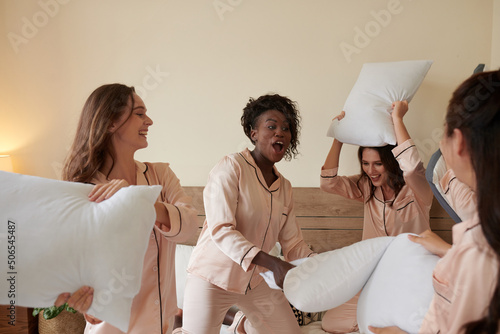 Group of excited young women in pajamas having pillow fight at sleepover party